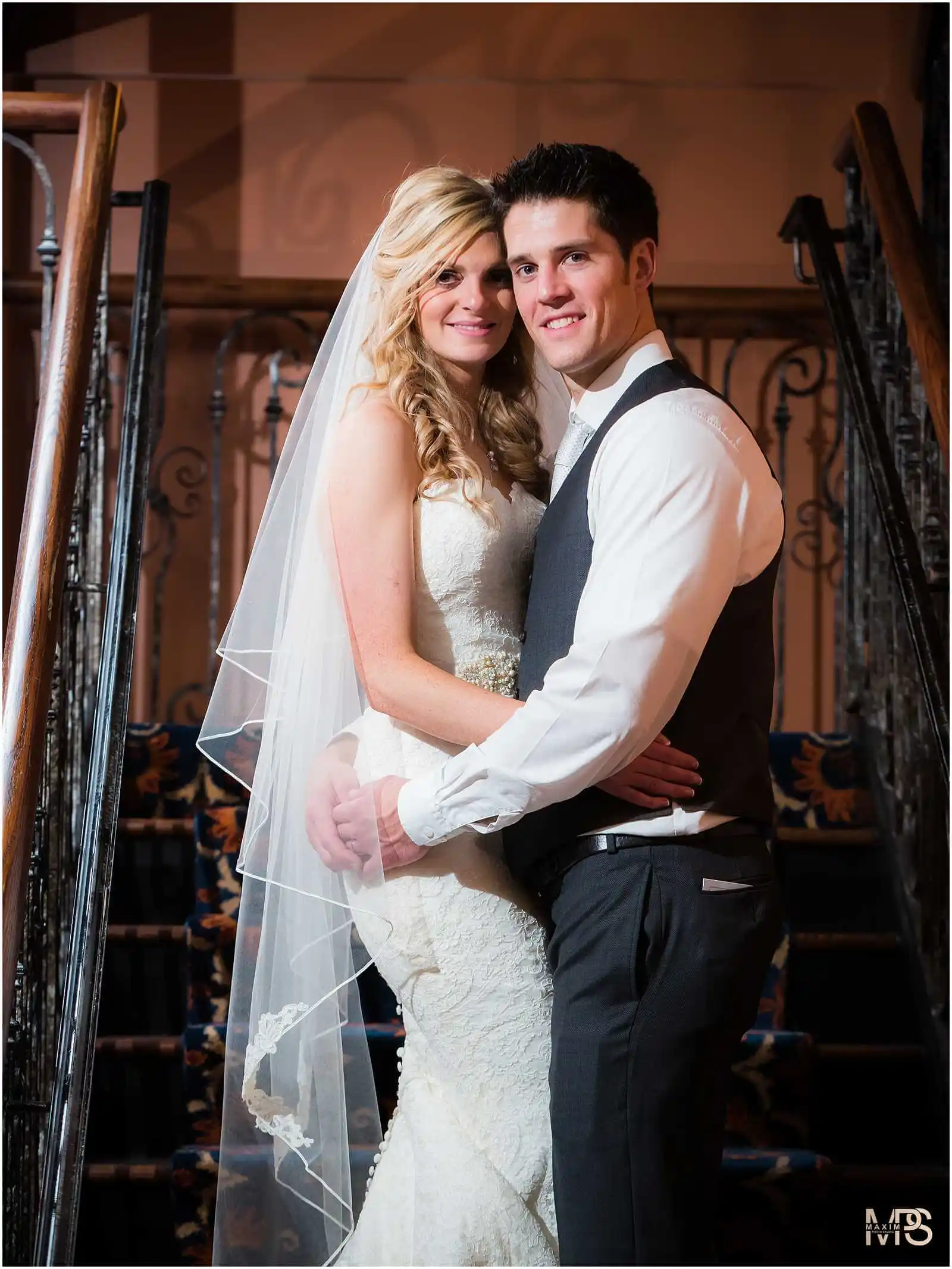Newlyweds embracing on grand staircase in wedding attire.