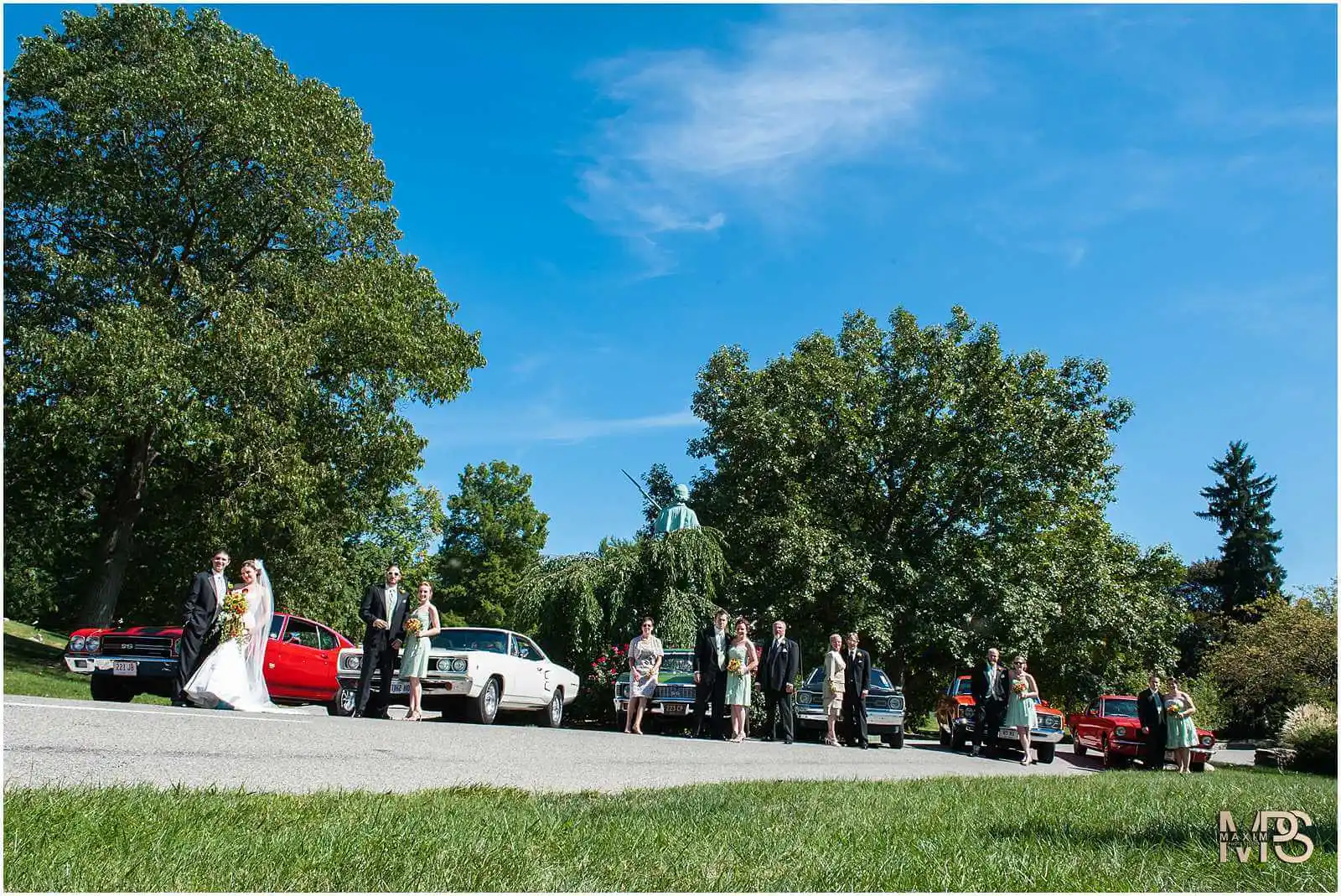 Outdoor wedding celebration with bridal party and vintage cars.